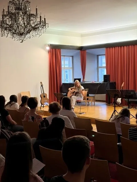 Eine Frau spielt Gitarre auf der Bühne eines Theaters. Ein Publikum sitzt vor ihr. Die Bühne ist mit roten Vorhängen dekoriert und beinhaltet Musikinstrumente.