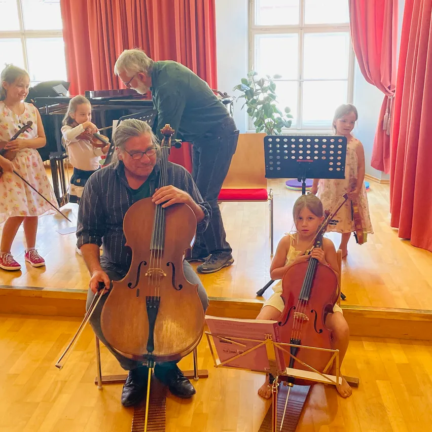 Ein Mann und zwei junge Mädchen spielen Cellos, während ein anderes Kind Geige in einem Raum mit Klavier und Notenständer spielt.