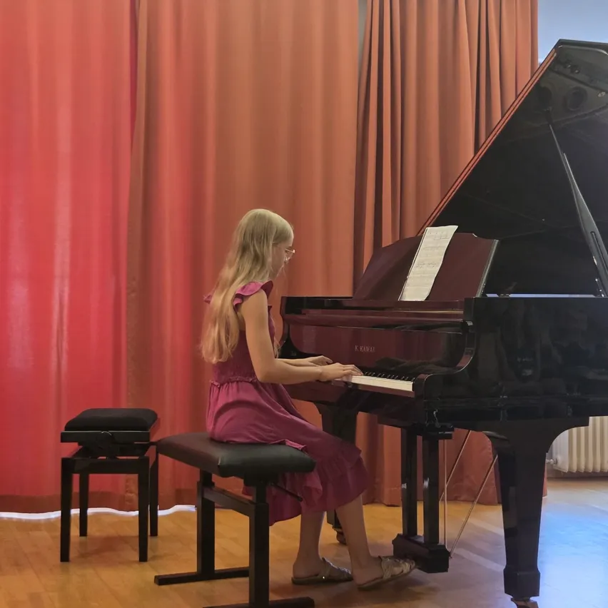 Ein junges Mädchen mit Brille spielt Klavier, auf einem Stuhl mit einem Notenständer davor sitzend. Das Klavier ist schwarz und im Hintergrund befindet sich ein roter Vorhang.