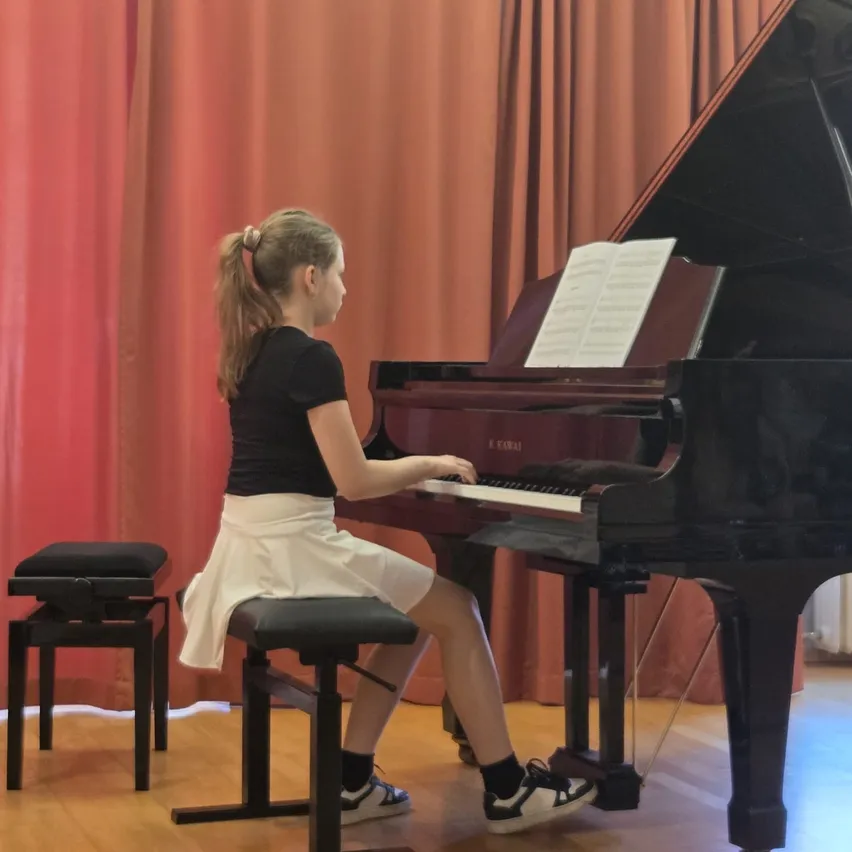 Ein Mädchen sitzt auf einem Klavierhocker und spielt Klavier, wobei Noten auf dem Klavier zu sehen sind. Das Klavier ist schwarz mit einer weißen Tastatur. Das Mädchen trägt ein schwarzes T-Shirt und einen weißen Rock.