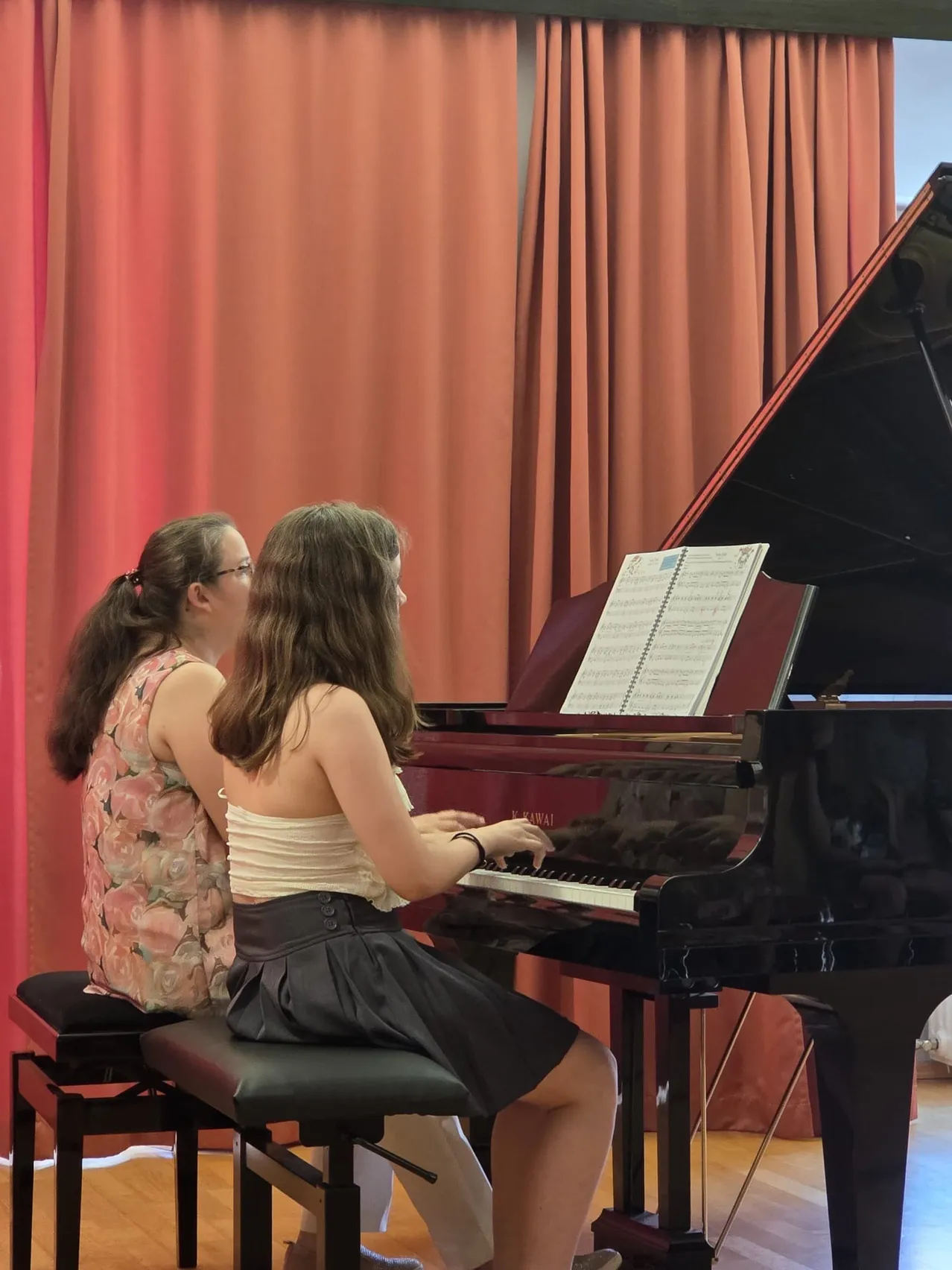 Zwei junge Mädchen sitzen an einem Flügel. Eine spielt, während die andere genau zusieht. Das Klavier hat ein offenes Musikbuch. Sie befinden sich in einem Raum mit roten Vorhängen.