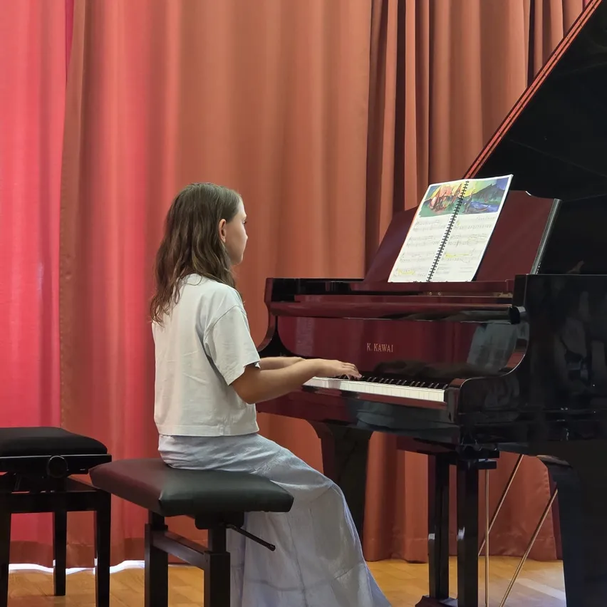 Ein junges Mädchen spielt Klavier mit einem Notenbuch vor sich. Sie trägt ein weißes T-Shirt und einen grauen Rock. Hinter ihr befindet sich ein roter Vorhang.