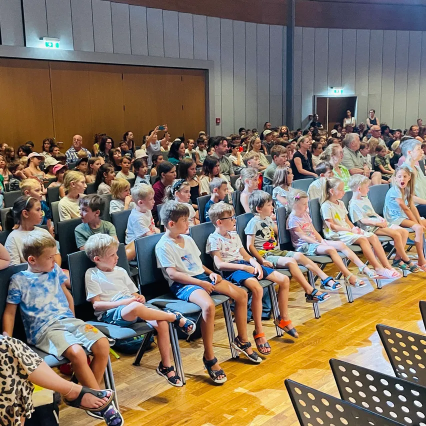 Eine große Gruppe von Kindern und Erwachsenen sitzt in Stühlen in einem Saal. Einige tragen Brillen und Sandalen. Der Saal hat Holzböden und weiße Wände.