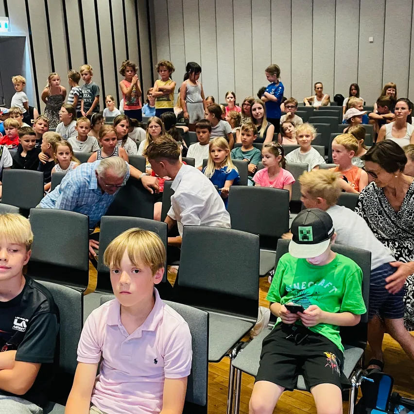 Ein Klassenzimmer voller Kinder. Einige sitzen, andere stehen hinter Stühlen und hören einem Erwachsenen vorne zu. Ein Junge mit einer Minecraft-Mütze schaut auf sein Handy.