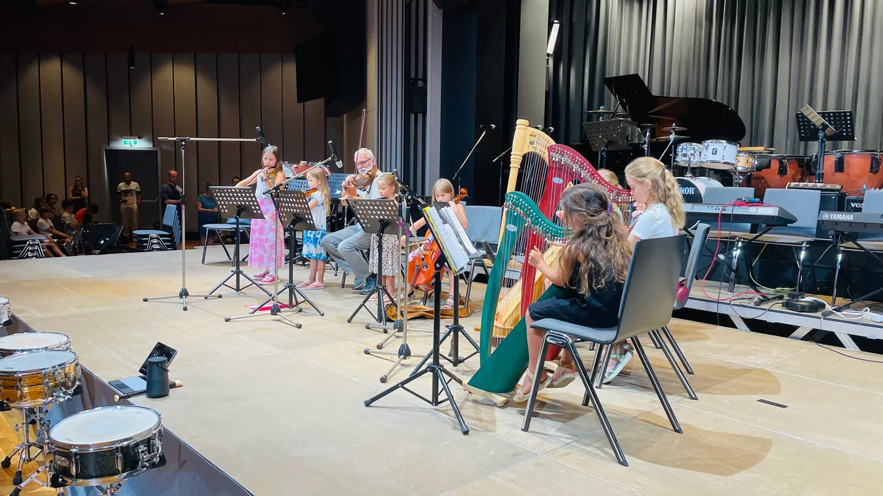 Eine Gruppe junger Mädchen und ein Mann spielen Instrumente auf der Bühne. Sie sitzen auf Stühlen und halten Violinen und Harfen. Der Mann steht und spielt die Violine. Hinter ihnen sind Stühle und ein Klavier.