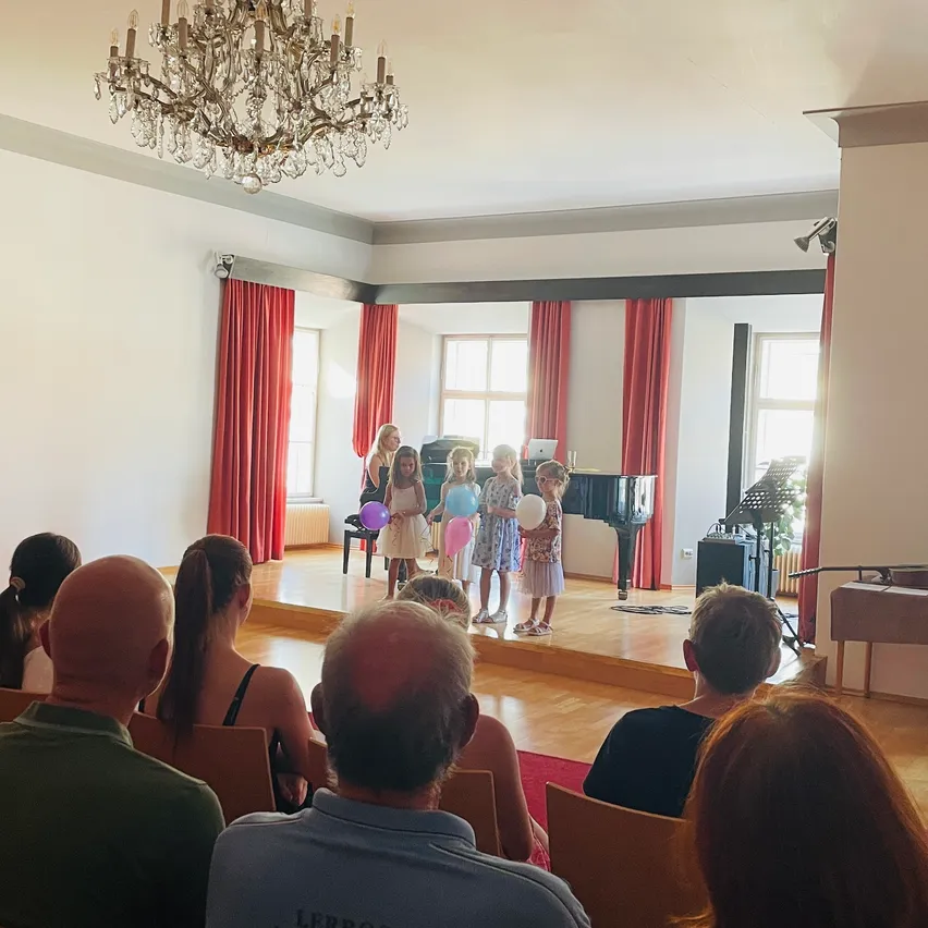 Eine Gruppe junger Mädchen mit Luftballons steht auf der Bühne, während einige Leute zuschauen. Ein Kronleuchter hängt von der Decke, und ein Klavier steht hinter den Mädchen.