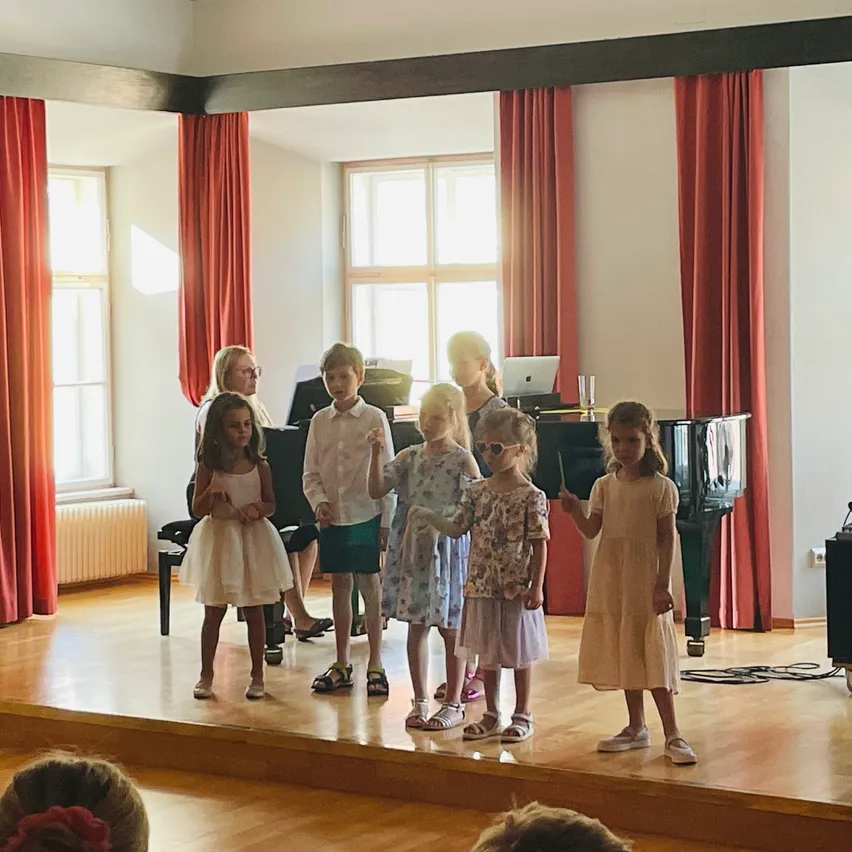 Mehrere Kinder in Kleidern stehen auf einer Bühne mit Klavier, roten Vorhängen und Fenstern. Sie scheinen eine Aufführung zu üben.