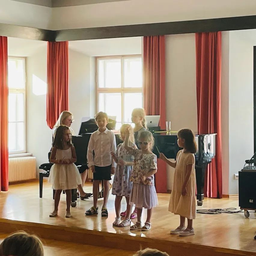 Eine Gruppe von Kindern steht auf der Bühne in einem Veranstaltungssaal mit einem Klavier im Hintergrund. Zwei Frauen stehen hinter den Kindern, eine mit einem Laptop. Der Raum ist gut beleuchtet mit roten Vorhängen.