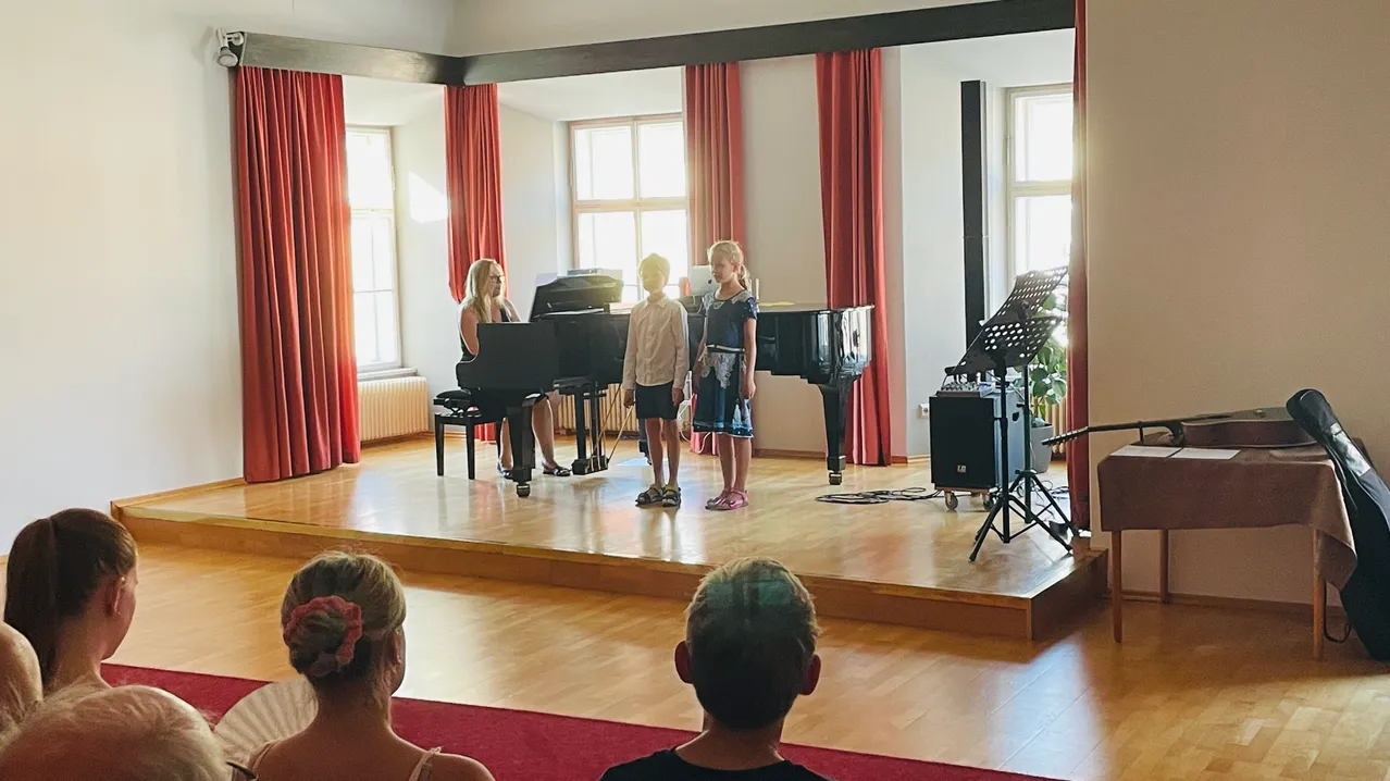 Eine Pianistin spielt auf der Bühne, während zwei Kinder vor ihr stehen. Ein Stativ mit Mikrofon steht auf der Bühne und ein Lautsprecher auf dem Boden. Eine Frau und ein Junge sitzen im Publikum.