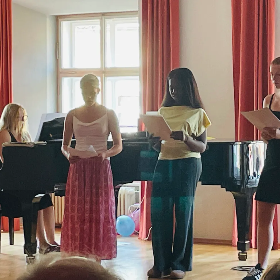 Eine Gruppe von Frauen steht in einem Raum mit einem Klavier und hält Papiere in den Händen. Zwei von ihnen lesen, während eine andere am Klavier sitzt. Ein Luftballon liegt auf dem Boden.