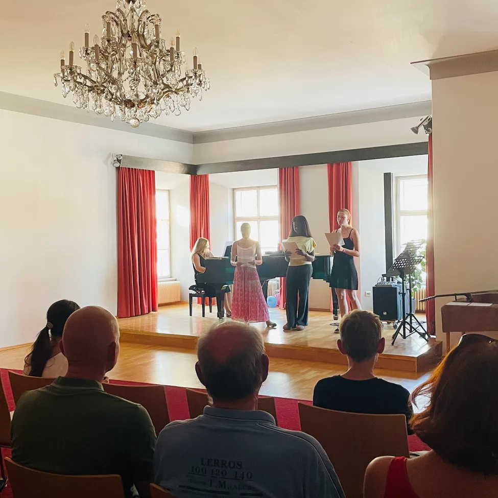 Eine Gruppe von Frauen steht mit Skripten auf der Bühne, während ein Klavier und ein Lautsprecher im Hintergrund zu sehen sind. Ein Publikum sitzt vor ihnen auf Stühlen.