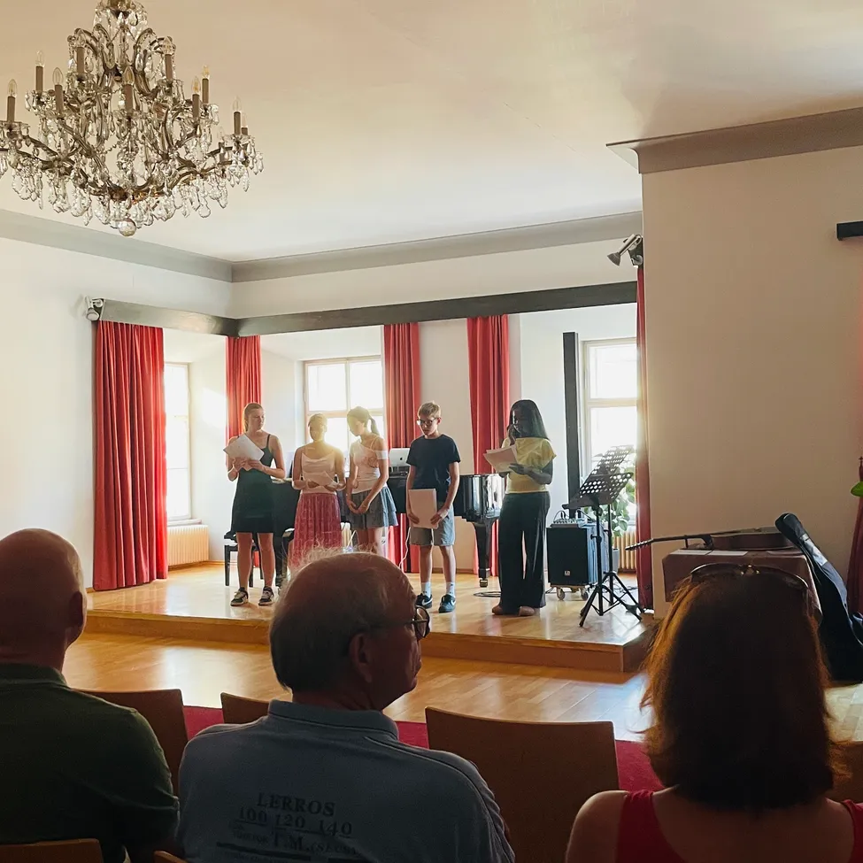 Eine Gruppe von Kindern steht auf der Bühne, hält Papiere in der Hand und sieht das Publikum an. Dahinter steht ein Klavier. Die Bühne ist mit roten Vorhängen dekoriert, und eine Kronleuchter hängt von der Decke.