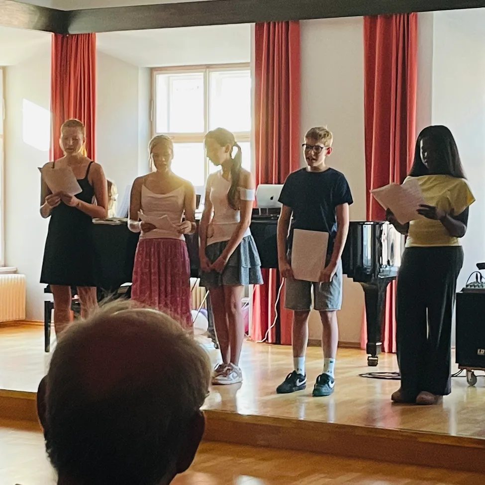 Fünf Schüler stehen auf einer Bühne und halten Papiere in der Hand, lässig gekleidet. Hinter ihnen ist ein Klavier und rote Vorhänge zu sehen. Der Boden ist aus Holz.