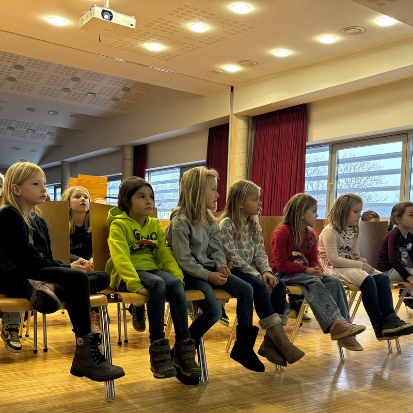 Mehrere junge Mädchen in verschiedenen Outfits sitzen aufmerksam in Stühlen in einem Raum, mit Blick nach vorne. Der Raum hat einen Projektor an der Decke und Fenster mit roten Vorhängen.
