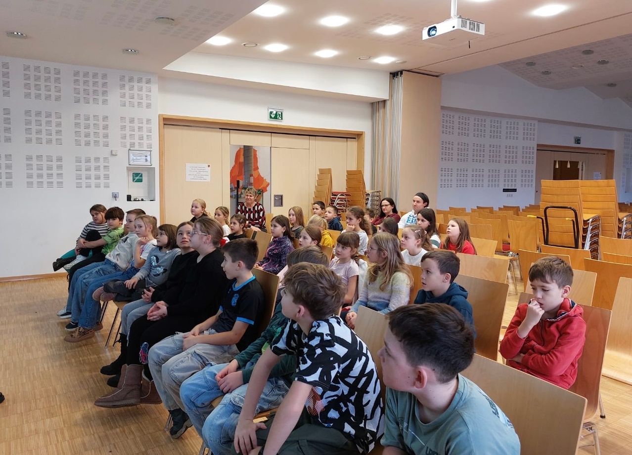 Eine Gruppe von Kindern sitzt in Reihen von Stühlen in einem großen Raum, nach vorne gerichtet. Der Raum hat Holzböden, weiße Wände und mehrere Deckenleuchten. Im Hintergrund befindet sich ein Notausgang und ein Projektor.