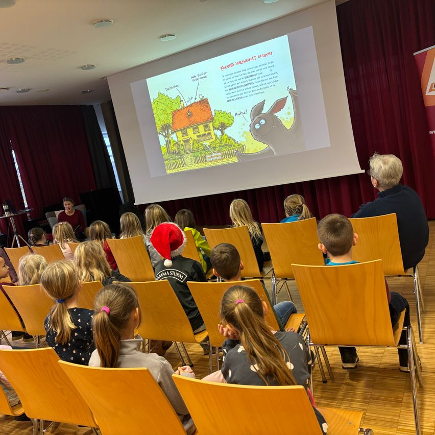 Ein Klassenzimmer mit Kindern in Stühlen, die einen Projektorbildschirm mit einer animierten Abbildung betrachten. Ein Mann mit einem Weihnachtsmannhut ist unter den Kindern.