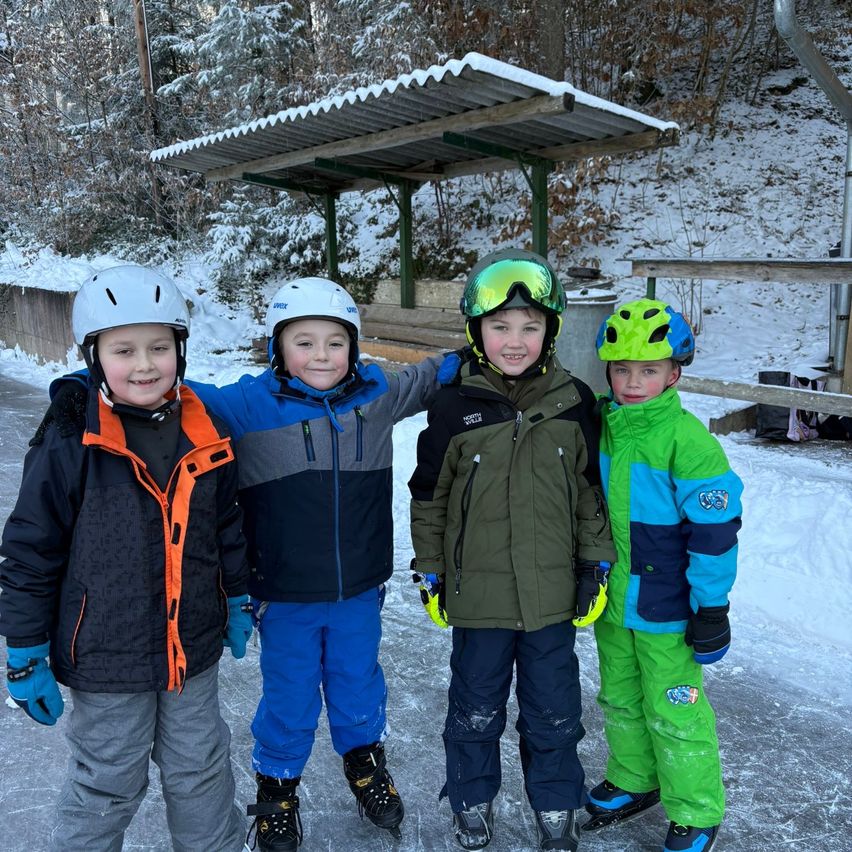Vier junge Jungen in Winterkleidung und Helmen posieren für ein Foto auf einer eisigen Oberfläche. Hinter ihnen ist ein schneebedeckter Hügel und ein Holzunterstand zu sehen.