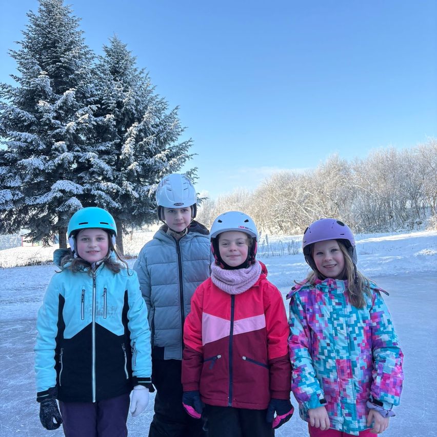 Vier Kinder stehen auf einer verschneiten Oberfläche, gekleidet in Winterkleidung. Drei Mädchen tragen Helme, zwei in Blau und eines in Lila. Ein Junge steht zwischen zwei Mädchen in einer grauen Jacke. Im Hintergrund sind schneebedeckte Bäume und ein klarer blauer Himmel zu sehen.