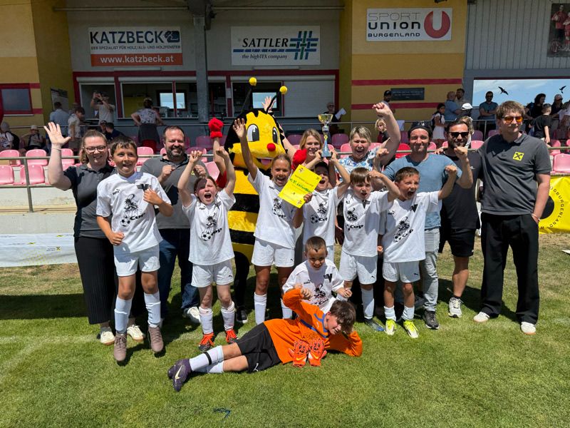 Eine Gruppe von Kindern und Erwachsenen posiert für ein Foto auf einem Fußballfeld. Sie tragen alle weiße Uniformen und halten einen Pokal. Der Maskottchen hält auch einen Pokal.