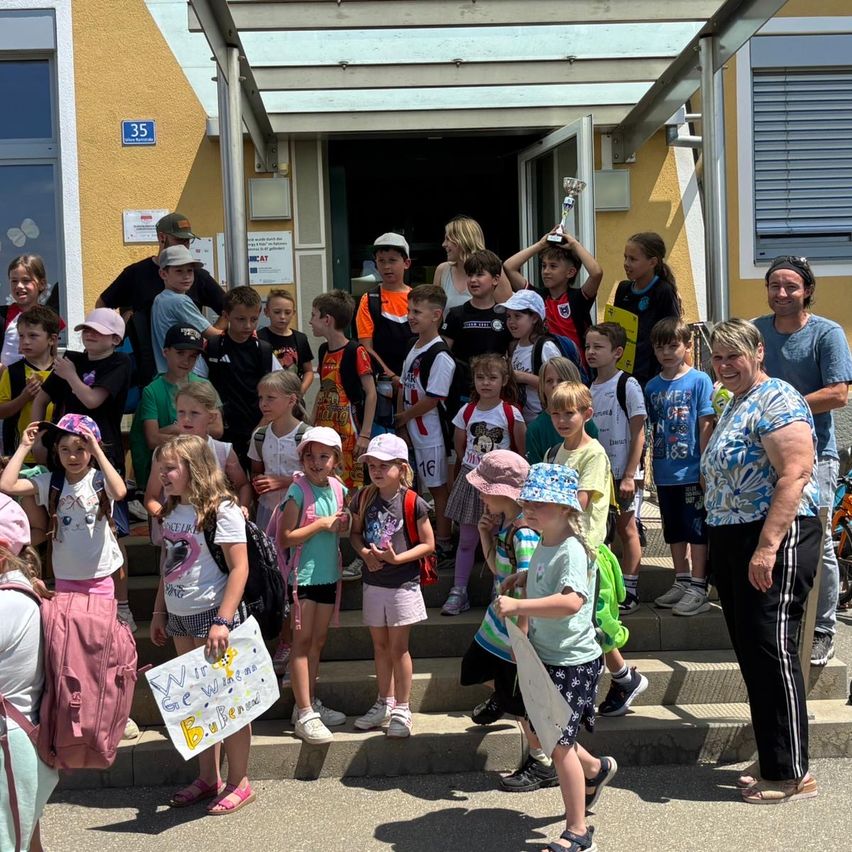 Eine Gruppe von Kindern, die Mützen tragen und Rucksäcke auf dem Rücken haben, steht auf Stufen vor einem Gebäude. Erwachsene stehen in der Nähe und lächeln. Ein Kind hält ein Schild. Das Gebäude hat eine Glastür und Fenster.