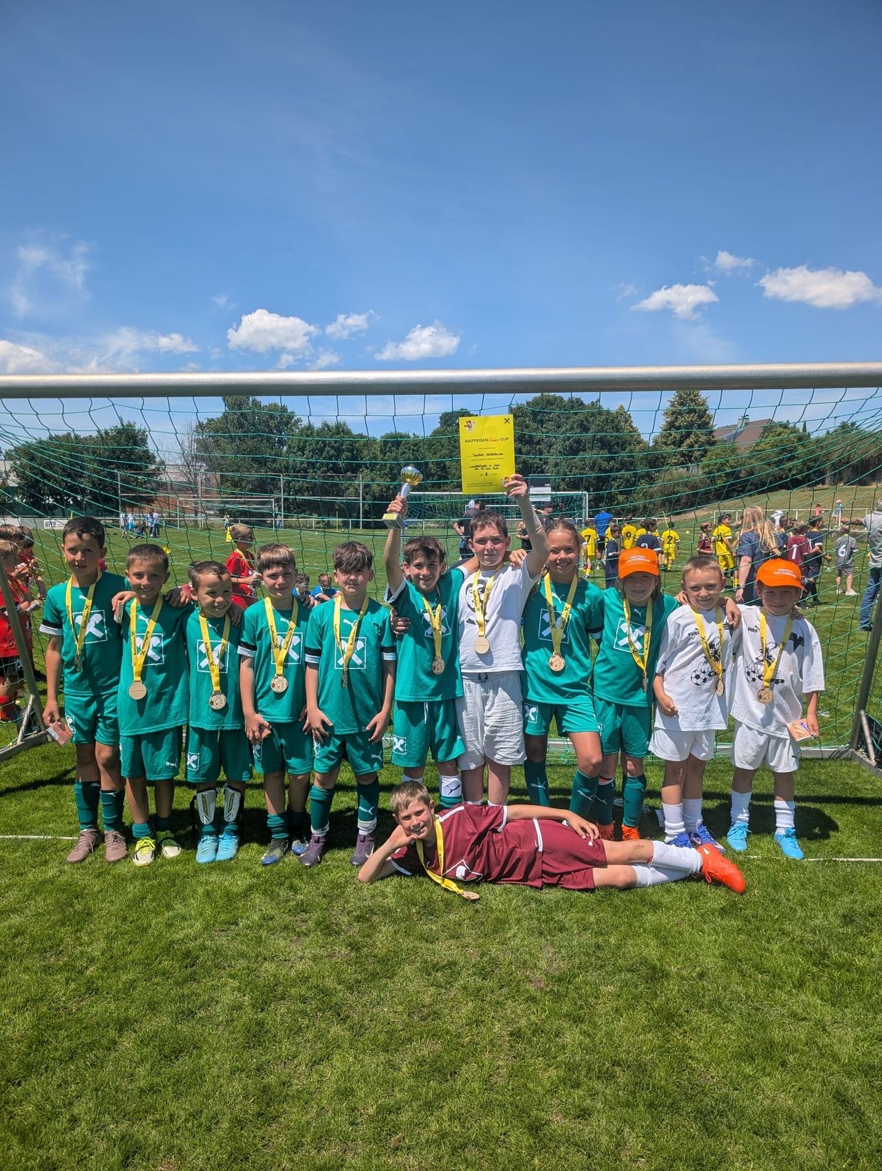 Eine Jugendfußballmannschaft steht vor einem Tornetz, alle tragen grüne Trikots und Medaillen, eine Mädchen hält einen Pokal.