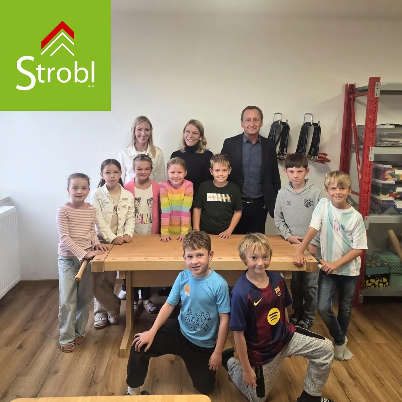 Eine Gruppe von Kindern und Erwachsenen posiert für ein Foto in einem Raum. Die Kinder stehen hinter einem Holztisch, und die Erwachsenen stehen hinter ihnen.