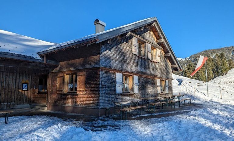 Ein Holzchalet mit Schornstein auf einem verschneiten Hügel, ein Fahnenmast mit einer Flagge in der Nähe und Picknicktische draußen.