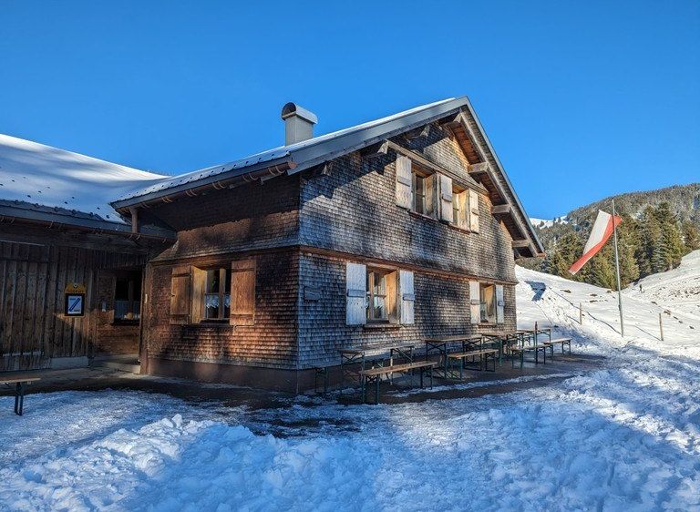 Ein Holzchalet mit Schornstein auf einem verschneiten Hügel, ein Fahnenmast mit einer Flagge in der Nähe und Picknicktische draußen.