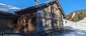 Ein Holzchalet mit Schornstein auf einem verschneiten Hügel, ein Fahnenmast mit einer Flagge in der Nähe und Picknicktische draußen.