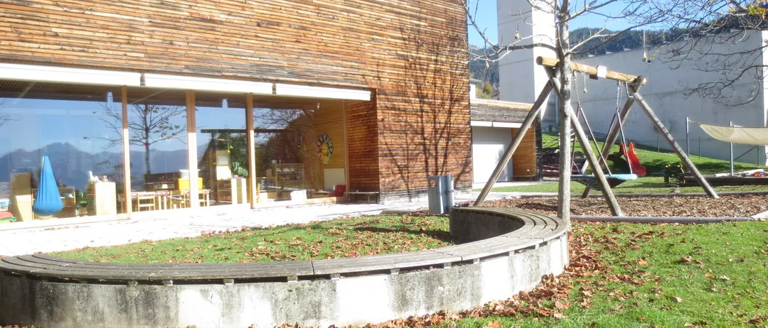 Ein modernes Gebäude mit einer Holzfassade und Glasfenstern. Draußen befindet sich ein Spielplatz mit Schaukeln und einer Bank. Der Boden ist mit Gras und abgefallenen Blättern bedeckt. In der Ferne sind Bäume und ein Gebäude zu sehen.