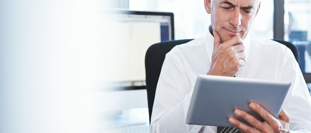 Ein älterer Mann in einem weißen Hemd und einer Krawatte sitzt an einem Schreibtisch, schaut auf ein Tablet und hat eine Hand am Kinn. Hinter ihm ist ein Monitor.