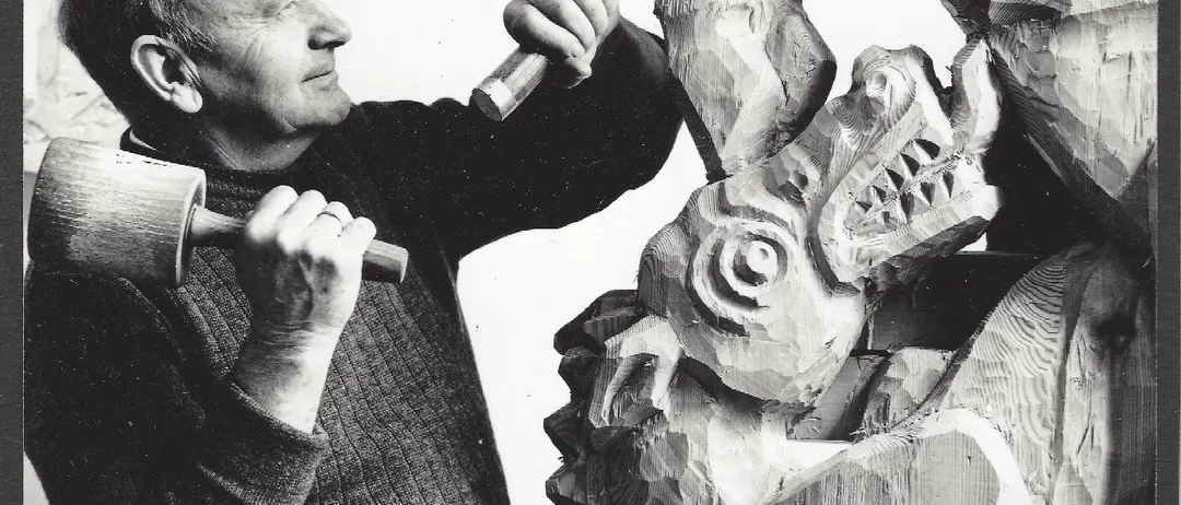Ein älterer Mann schnitzt eine Holzstatue einer Kreatur mit langer Schnauze und scharfen Zähnen. Er hält einen Hammer und Meißel in den Händen.