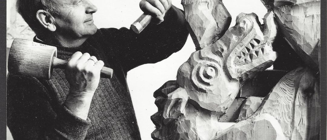 Ein älterer Mann schnitzt eine Holzstatue einer Kreatur mit langer Schnauze und scharfen Zähnen. Er hält einen Hammer und Meißel in den Händen.