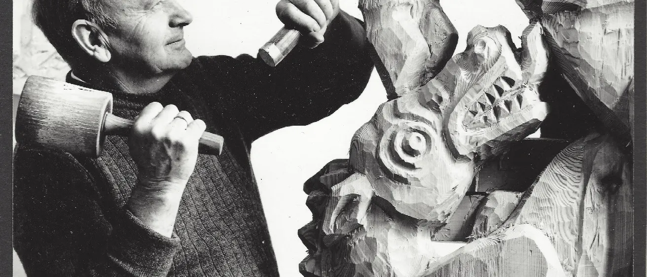 Ein älterer Mann schnitzt eine Holzstatue einer Kreatur mit langer Schnauze und scharfen Zähnen. Er hält einen Hammer und Meißel in den Händen.