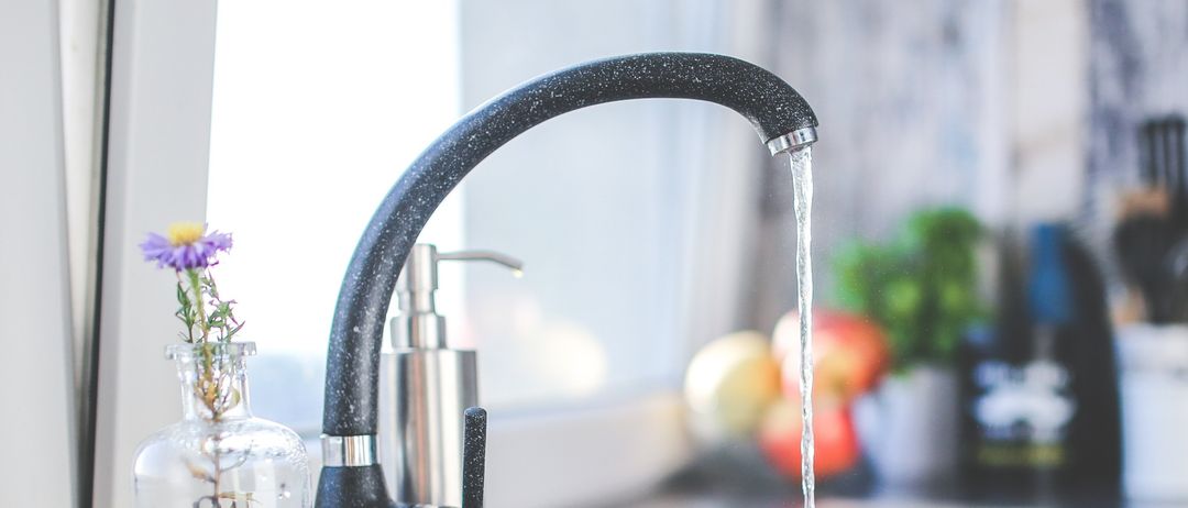 Eine Küchenspüle mit fließendem Wasser aus einem schwarzen Wasserhahn. Ein Glasvase mit Blumen und ein Seifenspender sind in der Nähe. Obst und Küchenutensilien im Hintergrund.