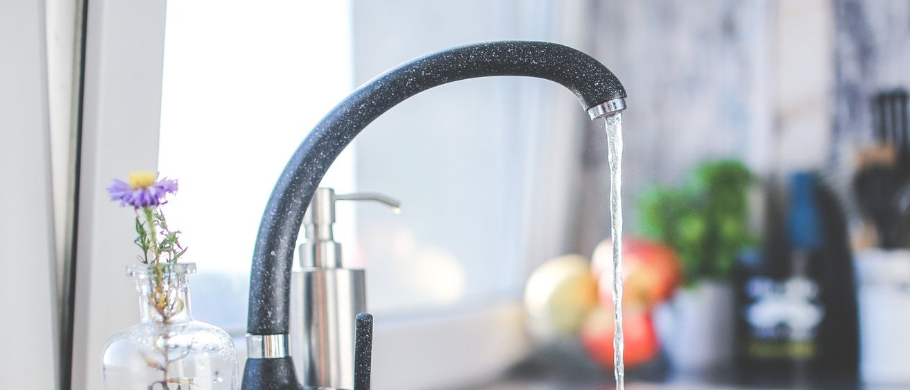Eine Küchenspüle mit fließendem Wasser aus einem schwarzen Wasserhahn. Ein Glasvase mit Blumen und ein Seifenspender sind in der Nähe. Obst und Küchenutensilien im Hintergrund.