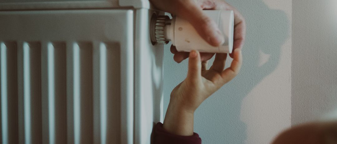 Ein Kind justiert den Thermostat an einem weißen Heizkörper mit der Hilfe eines Erwachsenen, vor einer hellblauen Wand.