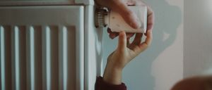 Ein Kind justiert den Thermostat an einem weißen Heizkörper mit der Hilfe eines Erwachsenen, vor einer hellblauen Wand.