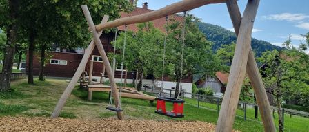Ein Spielplatz im Freien mit zwei Holzschaukeln, einem Haus im Hintergrund und Bäumen und Bergen in der Ferne.