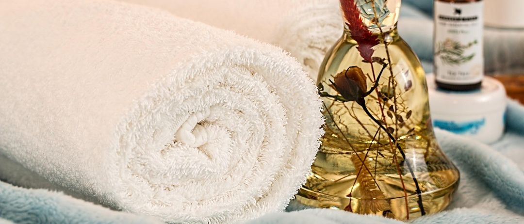 Zwei gerollte Handtücher und eine Flasche mit klarer Flüssigkeit und einer Blume darin auf einer blauen Decke.