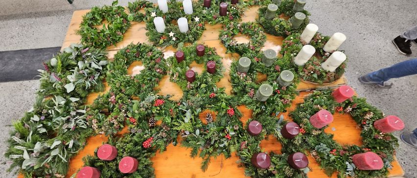 Ein Holztisch ist mit einer Reihe von Weihnachtskränzen gefüllt, die mit grünem Laub, roten Beeren und Kerzen in verschiedenen Farben geschmückt sind. Die Anordnung ist symmetrisch, wobei einige Kränze mit Tannenzapfen und Sternen verziert sind.