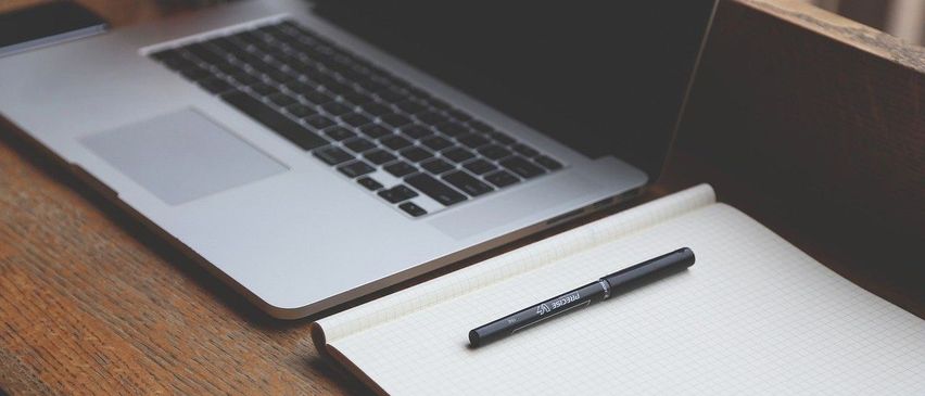 Ein silberner Laptop liegt auf einem Tisch mit einem Stift auf einem Notizbuch. Der Bildschirm des Laptops ist ausgeschaltet.