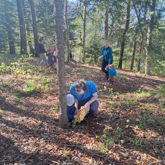 Ein Mann in einem blauen Hemd und mit Brille kniet sich hin und platziert einen gelben Beutel am Fuß eines Baumes. Ein kleines Kind in einem blauen Mantel und Hut steht in der Nähe. Im Hintergrund gehen eine Frau mit einem Kind und andere Personen auf einem Waldweg.