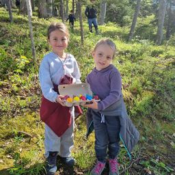 Zwei junge Mädchen stehen in einem Wald, eine hält eine Schachtel bunter Eier, während andere sich hinter ihnen versammeln.