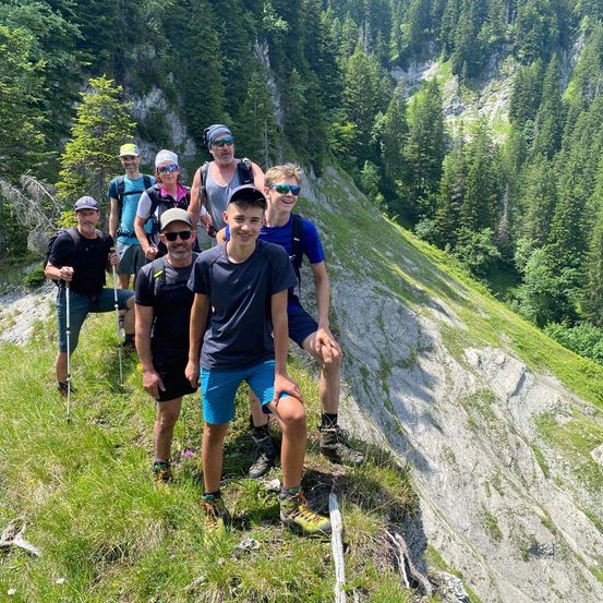 Eine Gruppe von Wanderern steht auf einem steilen, grasigen Berghang und trägt verschiedene Mützen und Sonnenbrillen. Sie stehen und posieren für ein Foto.