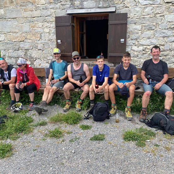 Eine Gruppe von Wanderern in Outdoor-Kleidung, die auf einer Bank vor einem Steingebäude sitzen, Flaschen halten und für die Kamera lächeln.