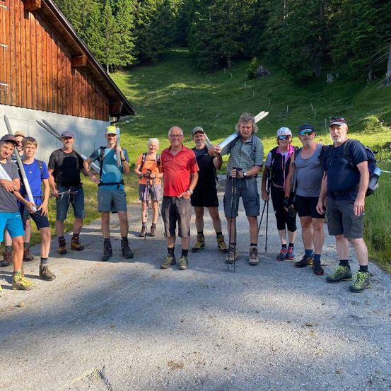 Eine Gruppe von Wanderern mit Trekkingstöcken steht vor einem Gebäude an einem sonnigen Tag, mit einem grün bewachsenen Hang im Hintergrund.