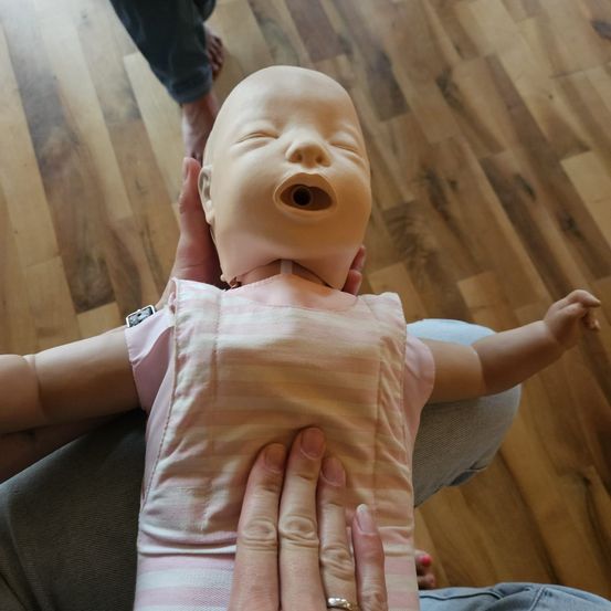 Eine Person hält eine Babypuppe mit offenem Mund auf einem Holzboden. Die Puppe trägt ein rosa Outfit mit Streifen. Eine weitere Person steht hinter der Person, die die Puppe hält.