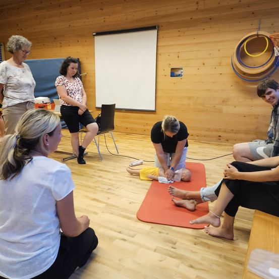 Mehrere Personen sitzen auf dem Boden und beobachten eine Frau, die auf einem roten Teppich auf einemDummy CPR durchführt. Eine Tafel ist an der Wand angebracht.