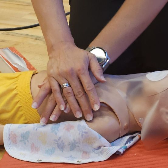 Eine Person in einem schwarzen Oberteil demonstriert Herz-Lungen-Wiederbelebung an einem Manikins, das ein gelbes T-Shirt trägt, mit Händen auf dessen Brust, wobei eine Uhr und ein Ring sichtbar sind.